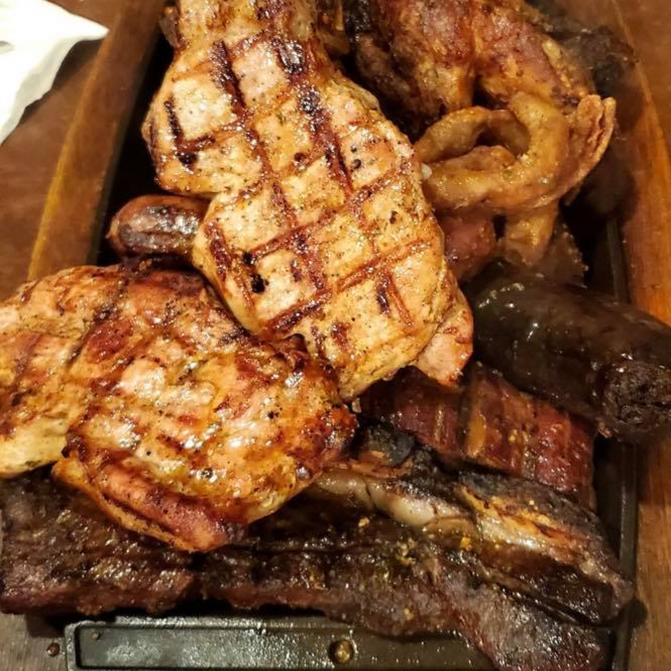 Close-up of a sizzling platter of various grilled meats, including steaks, sausages, and blood sausage.